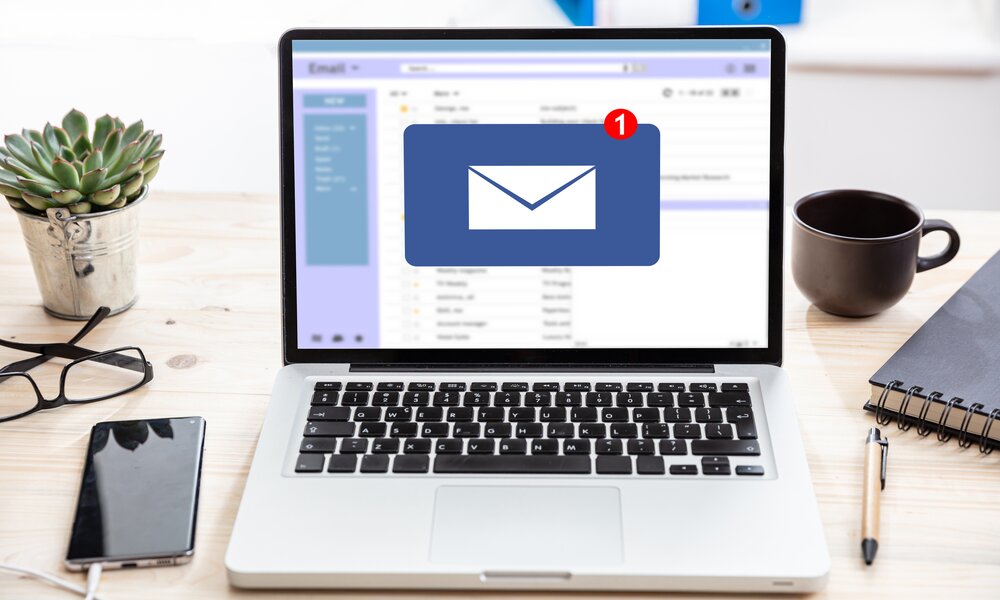 Ein aufgeklaptter Laptop auf einem Tisch, auf dem auch eine Tasse Kaffee, ein Smartphone, eine Brille und eine Pflanze zu sehen sind. Der Bildschirm des Laptop zeigt ein geöffnetes Email-Programm durch ein Briefsymbol an. | © Getty Images/Rawf8