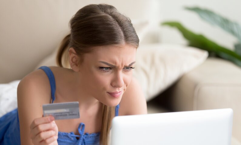 Junge Frau hält eine Kreditkarte in der Hand und verzieht das Gesicht, während sie auf den Bildschirm ihres Computers schaut. | © Getty Images/fizkes