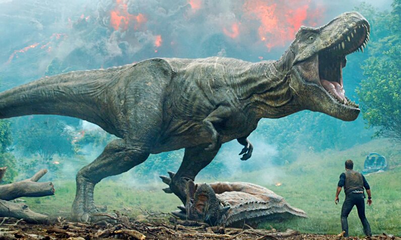Chris Pratt in einerSzene aus dem Film "Jurassic World: Das gefallene Königreich" - er steht als winzige Figur vor einem Tyrannosaurus Rex und Dschungelkulisse mit Feuer | © ddp images/LNKMEDIA/Universal Pictures 