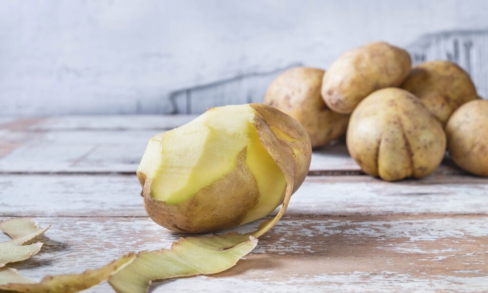 Eine halbgeschälte Kartoffel, im Hintergrund ein Haufen ungeschälter Kartoffeln | © Getty Images / Caterina Oltean / 500px