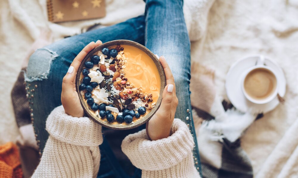 Eine Frau in Wollpullover und Jeans hält eine Schüssel mit Blaubeeren, Nüssen und Pudding in den Händen.  | © Getty Images / Xsandra
