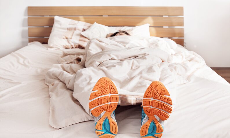 Eine junge Frau mit dunklen Haaren liegt im Bett und ihre Laufschuhe zeigen aus den Decken hervor. | © Getty Images / Solovyova